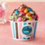 Milky Lane ice cream cup filled with Smarties and crushed Aero in front of a bowl of marshmallows on a pink surface.